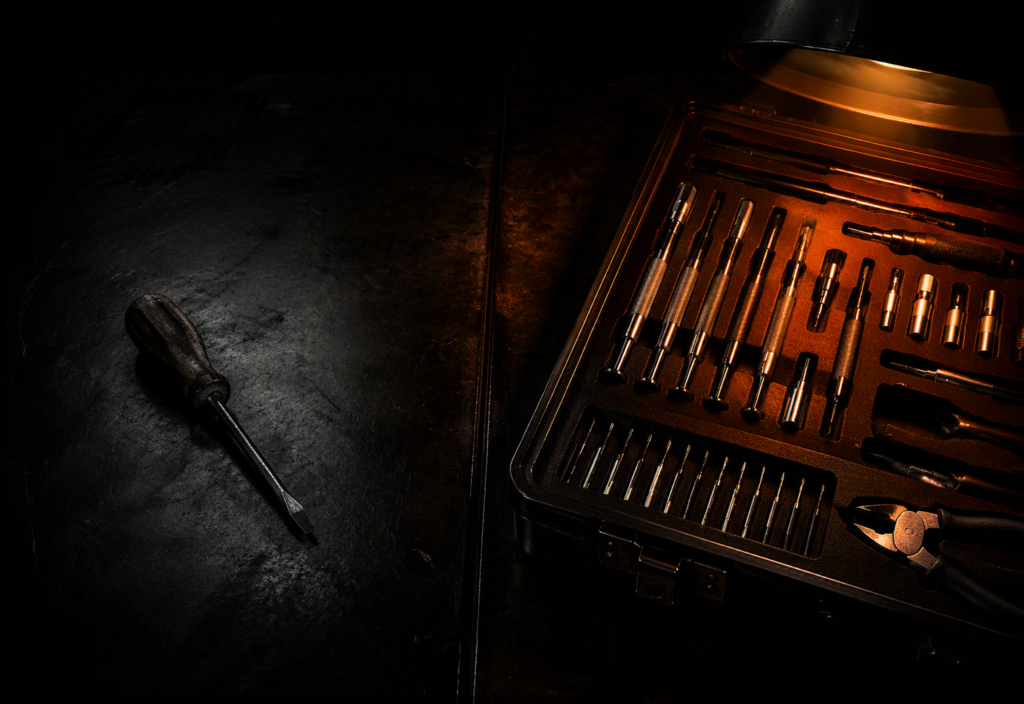 A dark industrial workbench split between a single worn screwdriver in shadow on the left and an open professional precision toolkit glowing under warm orange light on the right
