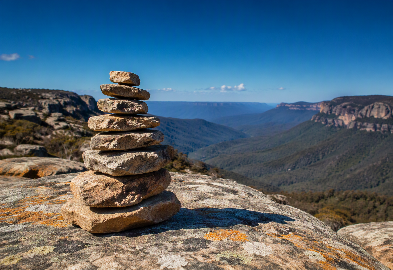 A stone cairn of five to seven carefully selected and stacked flat granite slabs stands alone on an exposed rocky Australian mountain summit, with a wide panoramic valley of layered eucalyptus ridgelines receding into blue haze below under a clear deep blue sky and warm directional sunlight. The cairn represents deliberate, verified documentation — each stone individually assessed and placed — while the vast landscape beyond communicates the scope of what can now be mapped and governed from a position of clarity.