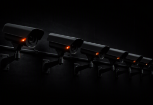 A row of industrial surveillance cameras in dark matte steel mounted on steel arms, each with a single warm amber-orange active indicator LED glowing against a near-black background
