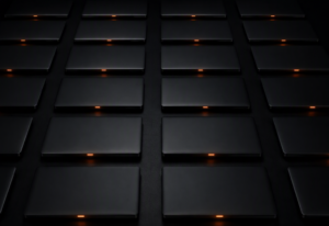 A regular grid of identical closed dark laptops on a flat near-black surface, each with a single small warm amber-orange indicator LED glowing on its lid