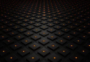 A regular grid of small dark matte square tiles filling an ultra-wide near-black surface, each tile with a single warm amber-orange pinpoint of active light at its centre, subtle brightness variation across the grid
