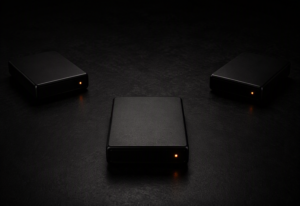 Three identical small dark matte sealed storage device enclosures arranged in a precise triangular formation on a near-black surface, each with a single warm amber-orange active indicator LED, physically separated with no cables connecting them