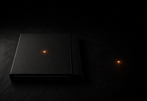 A sealed dark matte professional document folder lying flat on a near-black surface with an amber-orange indicator dot centred on its cover, and a second identical amber-orange dot resting alone on the bare surface at a deliberate distance from the folder